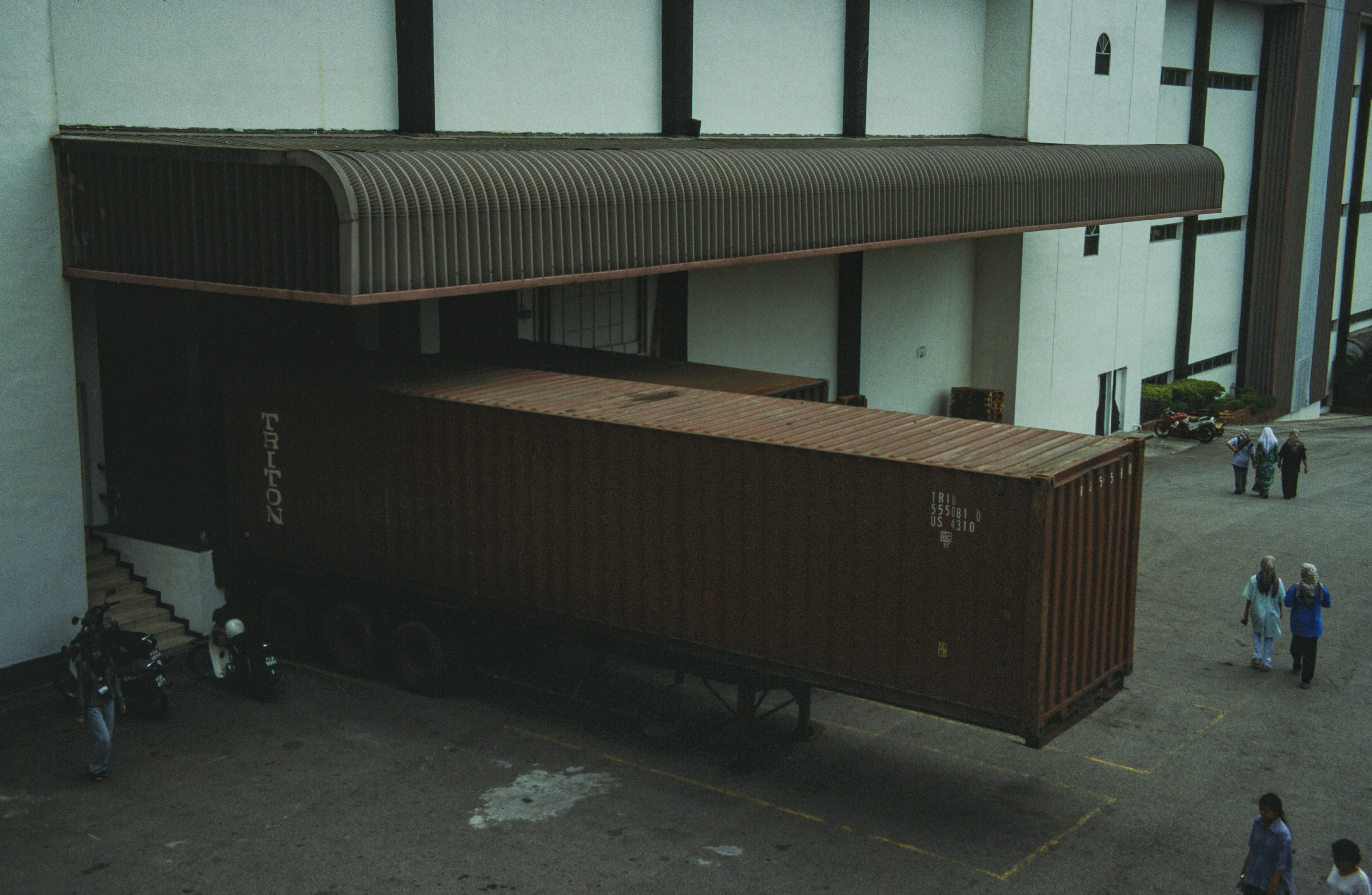 The image shows a large shipping container attached to a truck, backed into a loading dock at an industrial warehouse. The dock is covered by a curved awning, and several people and motorbikes are visible in the surrounding area. The scene depicts a typical logistical operation, with the container being prepared for loading or unloading at a commercial facility.